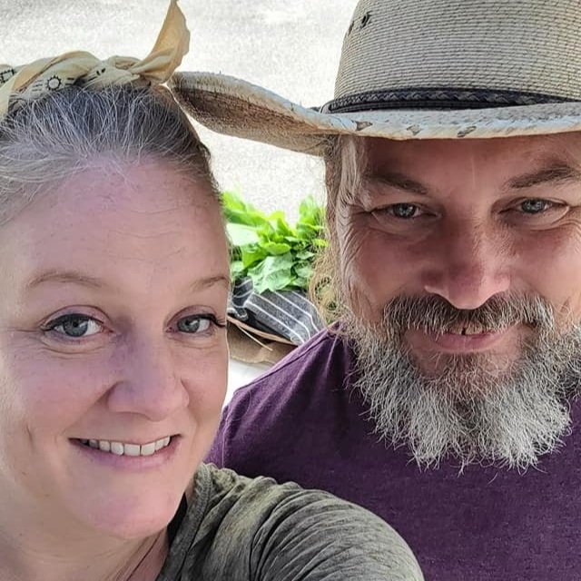 Smiling couple posing for a selfie outdoors, with a straw hat and casual clothing, surrounded by greenery and fresh produce in the background.